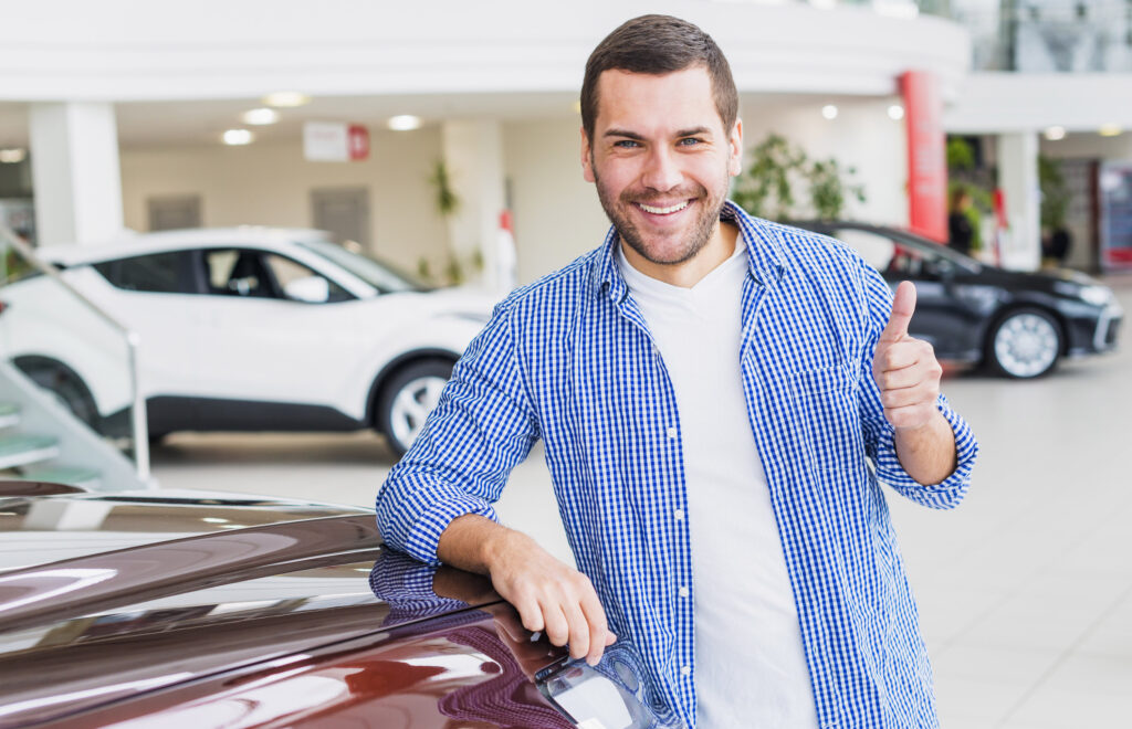 Homem sorrindo, vestindo camiseta branca e, por cima, camisa xadrez azul, fazendo sinal de positivo com o dedo. Ele se escora em um carro na cor vinho, que aparece muito pouco. Ao fundo, desfocado, aparecem dois outros carros, um preto e um branco, em um ambiente comercial