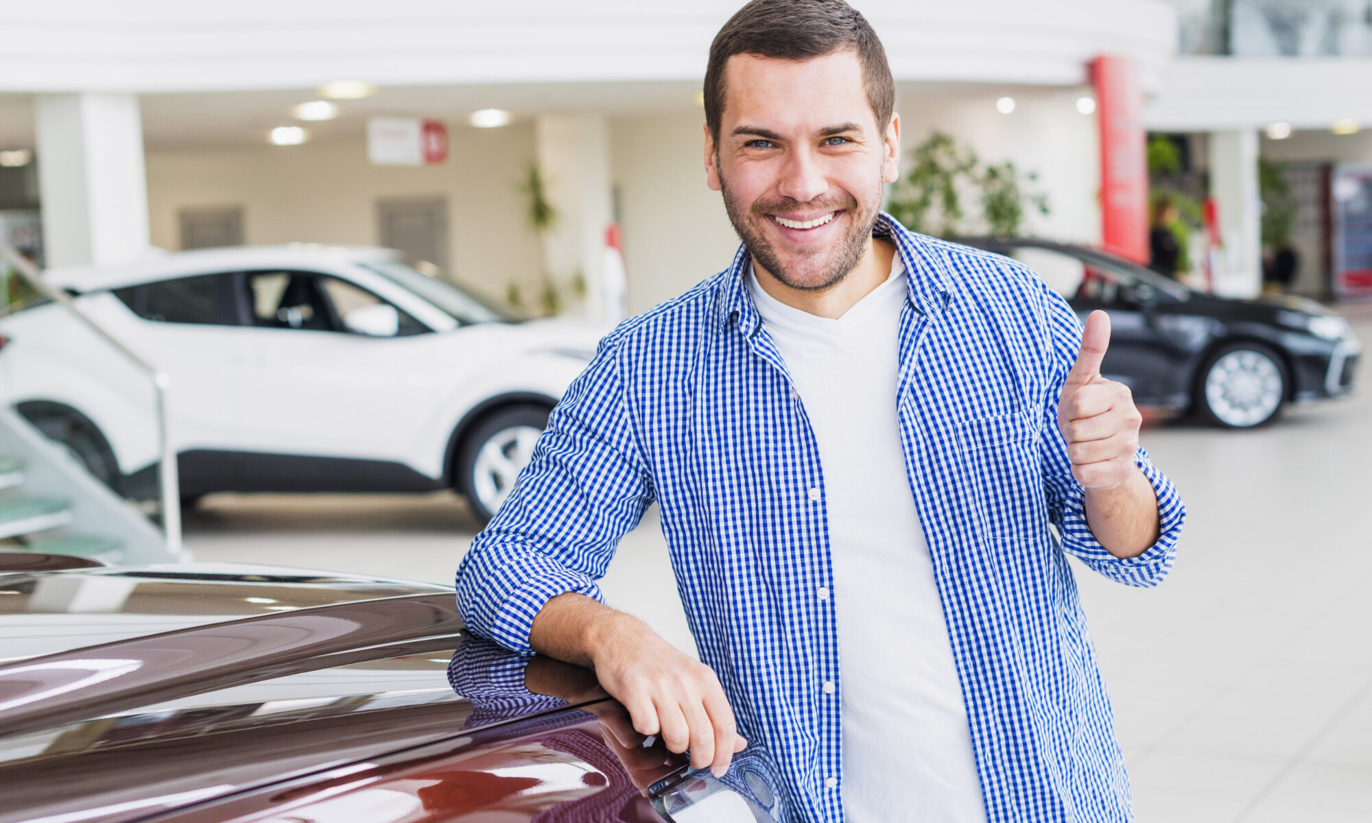 Homem sorrindo, vestindo camiseta branca e, por cima, camisa xadrez azul, fazendo sinal de positivo com o dedo. Ele se escora em um carro na cor vinho, que aparece muito pouco. Ao fundo, desfocado, aparecem dois outros carros, um preto e um branco, em um ambiente comercial